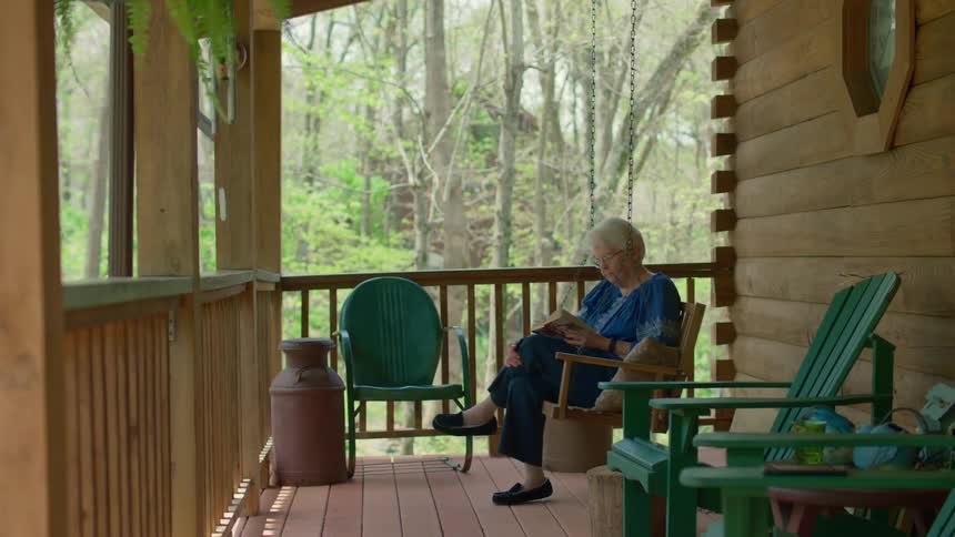 An elderly woman sits on her front porch outside and reads a book