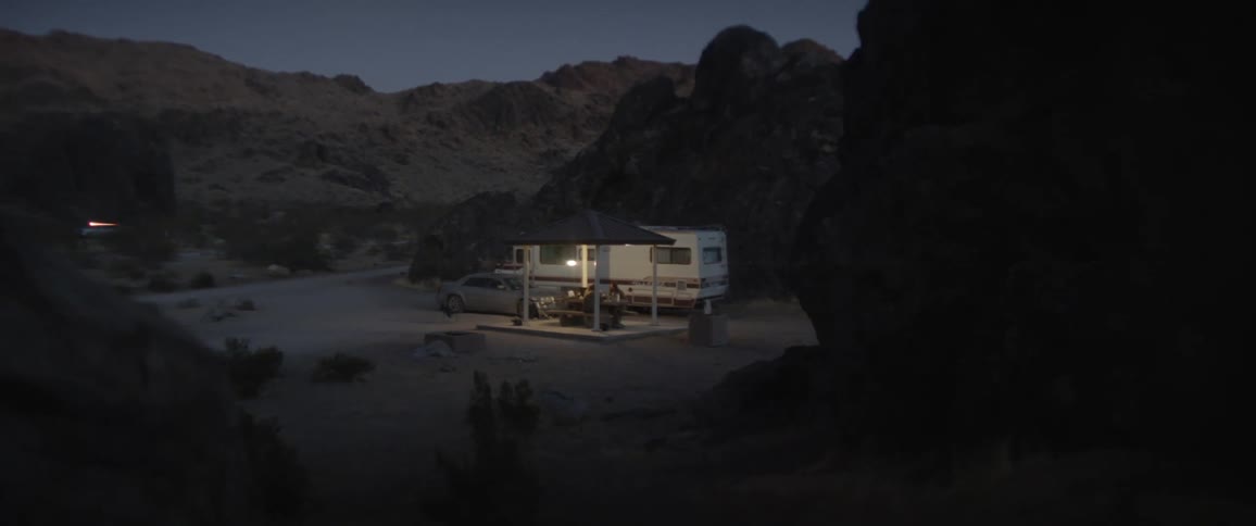 People sit under pavilion in arid area surrounded by hills near rv and car at night