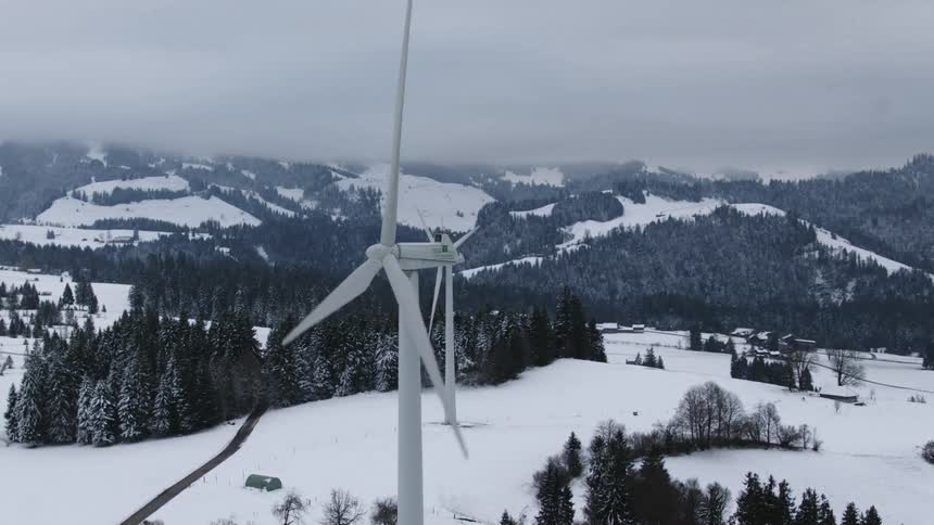 Wind turbines spin on a snow covered mountain surrounded by forested mountains under a hazy sky