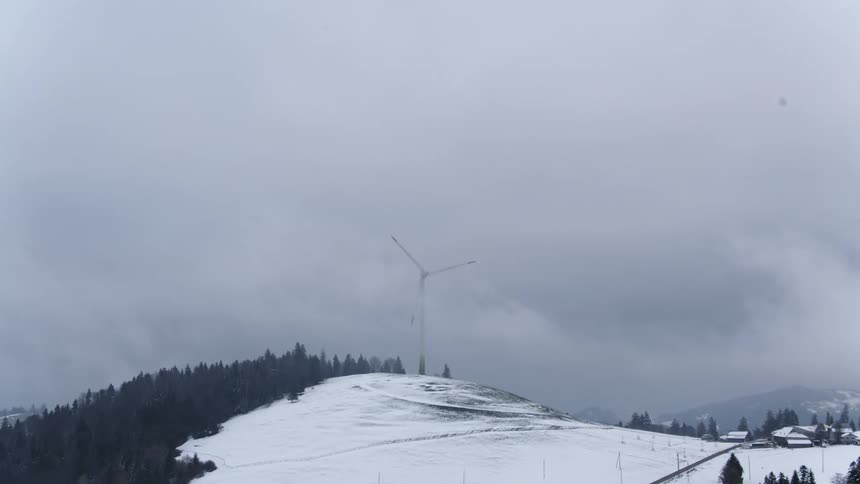 Wind turbine on snow and forest covered mountain towers over nearby cluster of buildings