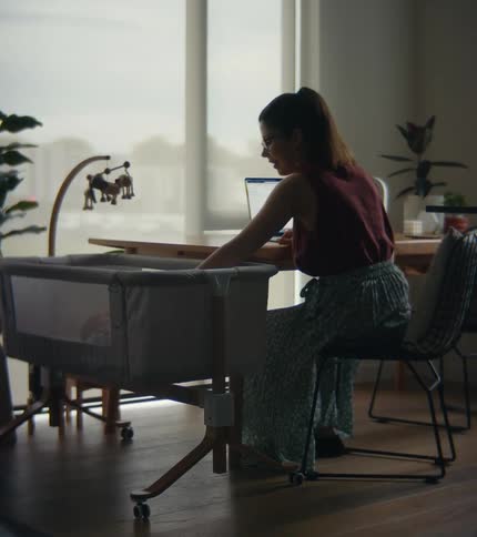 A woman is sitting at a table in front of windows with a laptop computer as she reaches into a playpen