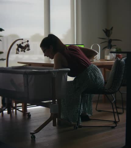 A woman is sitting at a table in front of windows with a laptop computer as she reaches into a playpen
