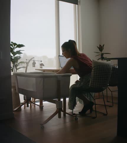 A woman at home sits in front of a laptop with a cradle next to her and reaches down to comfort a baby