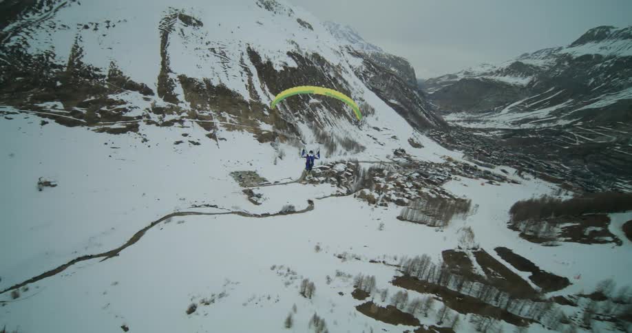 A paraglider wearing skis is flying along the side of mountains and over a valley