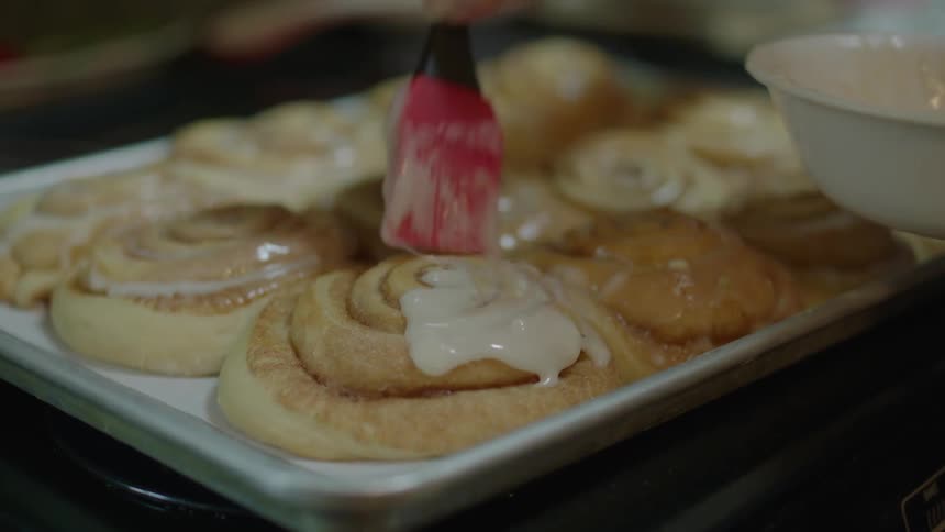 A person frosting cinnamon rolls with white frosting on a large pan