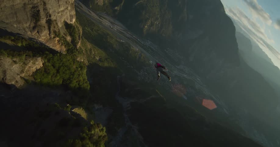 A skydiver with a parachute pack on their back is floating along the side of a mountain cliff and over the valley below