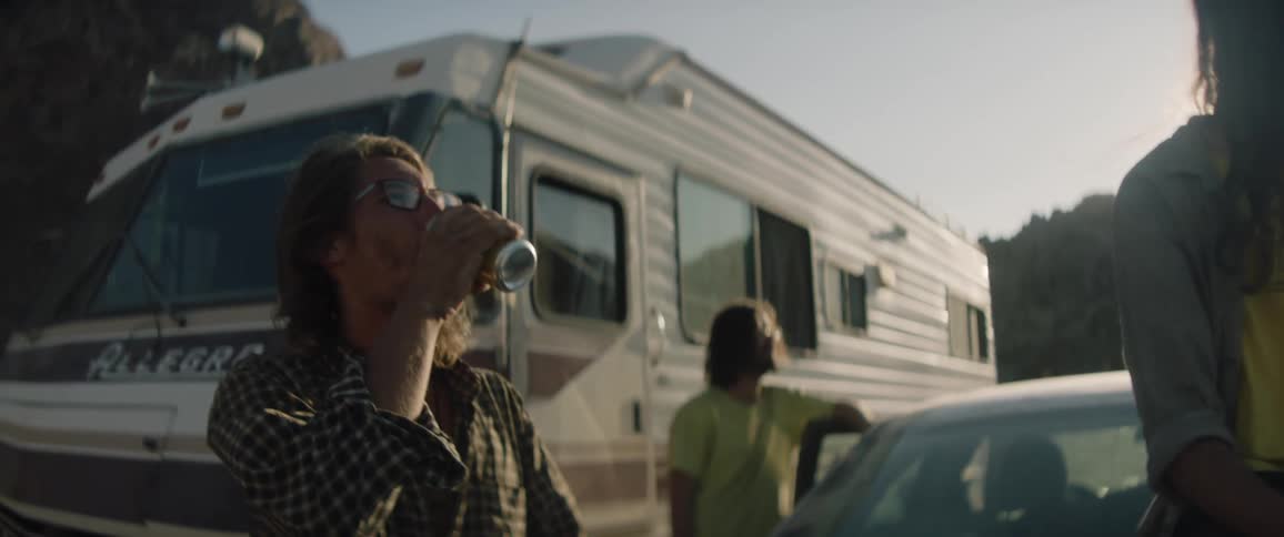 A man drinking from a can around vehicles and other people with the sun shining on them