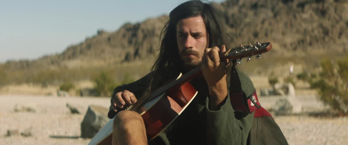 A man in front of a mountain range plays a guitar as another man walks at the base of the mountain