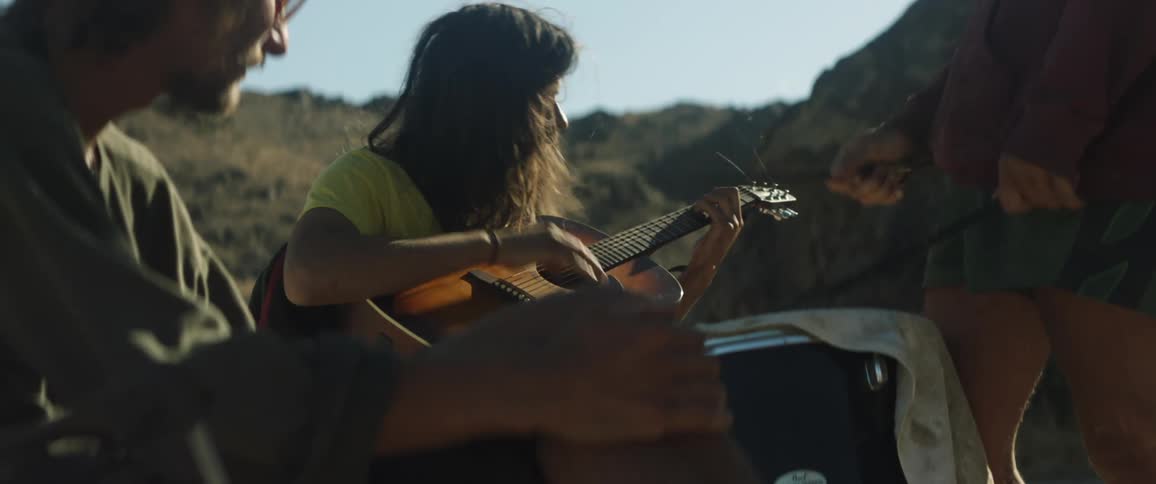 Three people are sitting together outside as a man plays a guitar and another person plays a drum