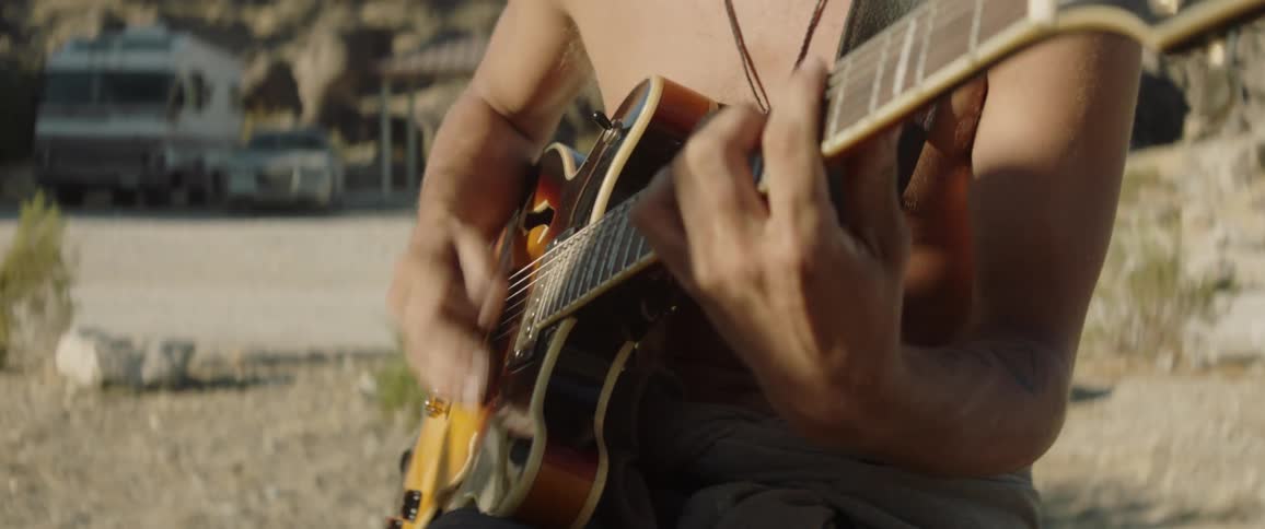 A shirtless man strums a guitar at a desert campsite