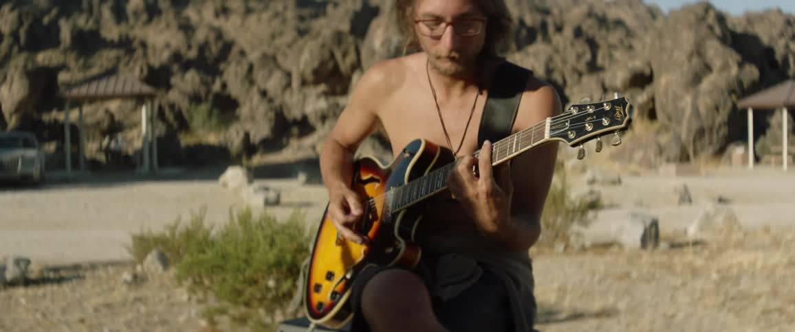A shirtless man playing an electric guitar outside on a sunny day