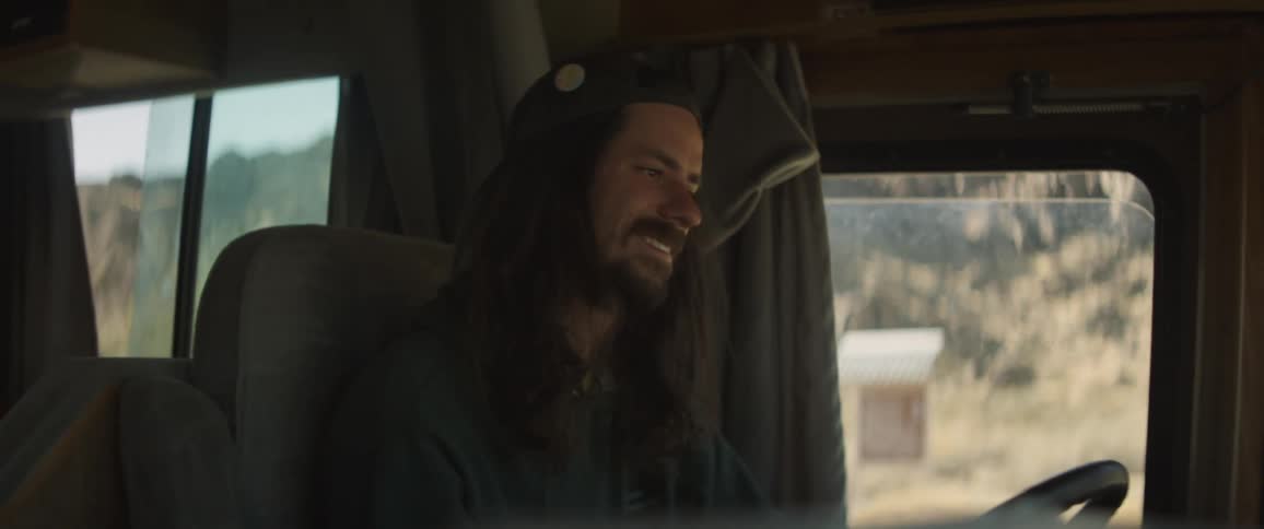 A man with long hair and a backwards baseball cap driving an rv through a desert mountainous area