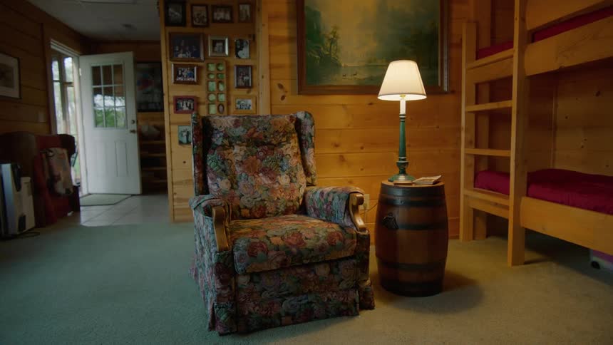 A lamp is on next to a chair and a bunk bed with a ladder as the front door is left open
