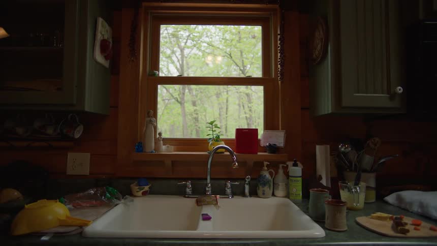 A small kitchen window with a white kitchen sink in front of it with different items like a strainer on the counter