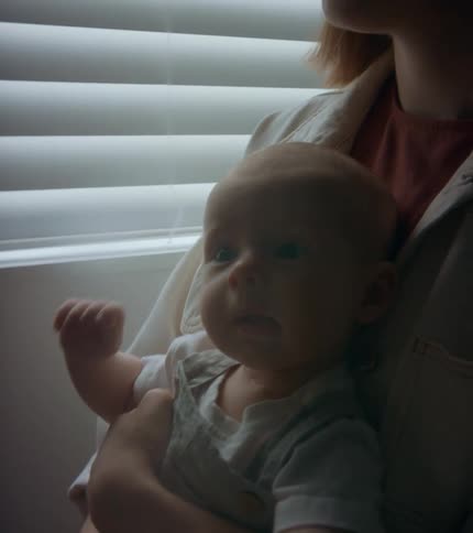 An infant squirming in a persons arms next to a window
