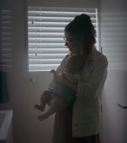 A mother holds her baby standing looking in the mirror next to a window