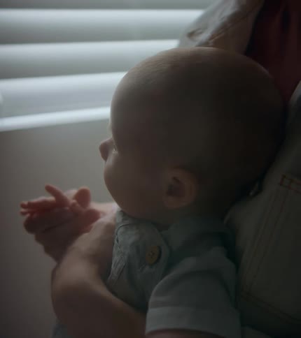 A woman is standing in a room near a window shade holding her baby in her arms and rubbing their hand