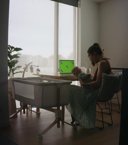 Mother with ponytail glasses and blue dress sits at table holding baby