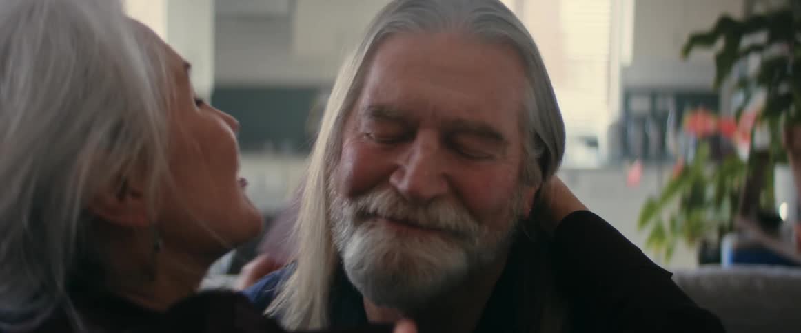 Woman with gray hair and brown shirt touches and kisses on elderly mans cheek