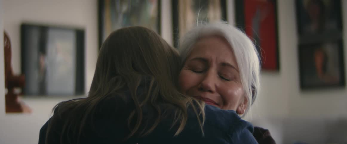 Elderly man with long gray hair and dark blue shirt cuddles woman with gray hair in room with pictures