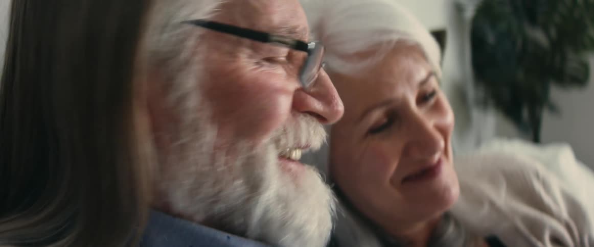 Elderly man with gray hair beard and glasses sits next to woman with gray hair in room laughing