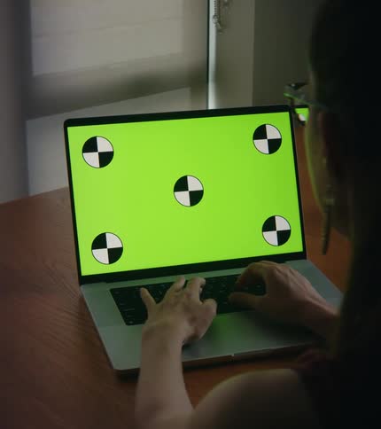 Woman sitting at a desk typing on the keyboard of a laptop with a replaceable green screen