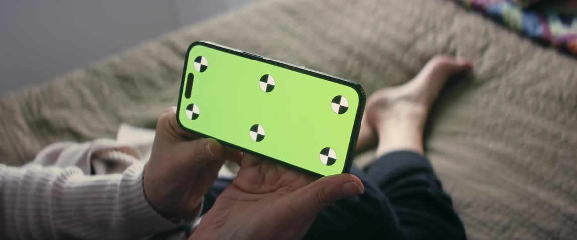 A person holds a smartphone while sitting on a bed indoors during the day