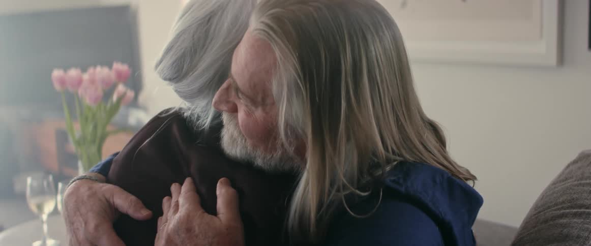 Elderly man with long gray hair and beard kisses and hugs old woman in living room at home