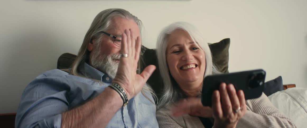 Elderly man and woman sitting on bed make video call on smartphone in room