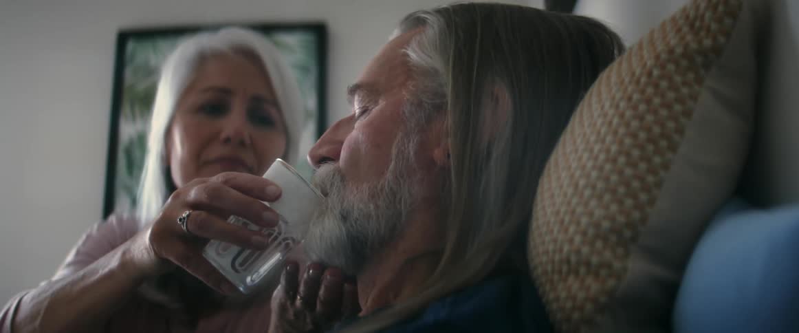 Woman with gray hair gives pill and glass of water to elderly man sitting on bed in room
