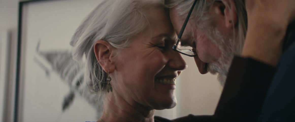 Elderly man with long gray hair glasses and beard and woman with gray hair and brown shirt dance happily