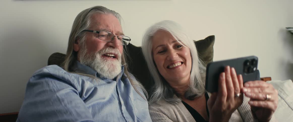 Old man with long gray hair beard glasses and shirt sits on bed next to woman with gray hair making video call on smartphone in room