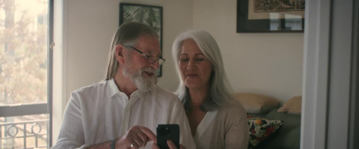 Two elderly people standing in the livingroom looking at a smartphone and laughing