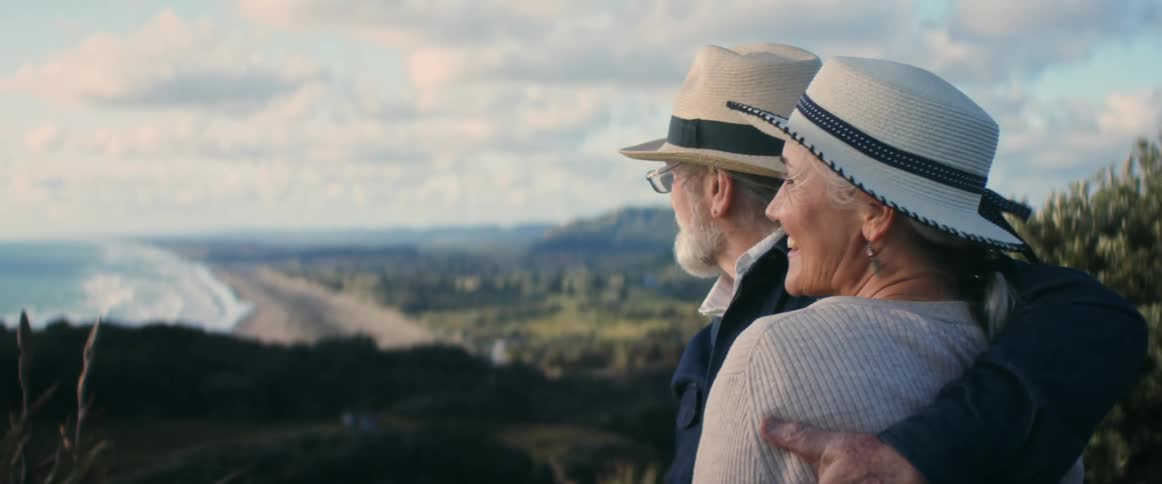Elderly man and woman wearing hats and jackets stand on mountain overlooking coast