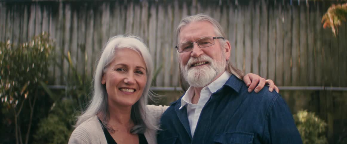 Elderly man with long hair and beard and woman with gray hair smile at camera in the yard