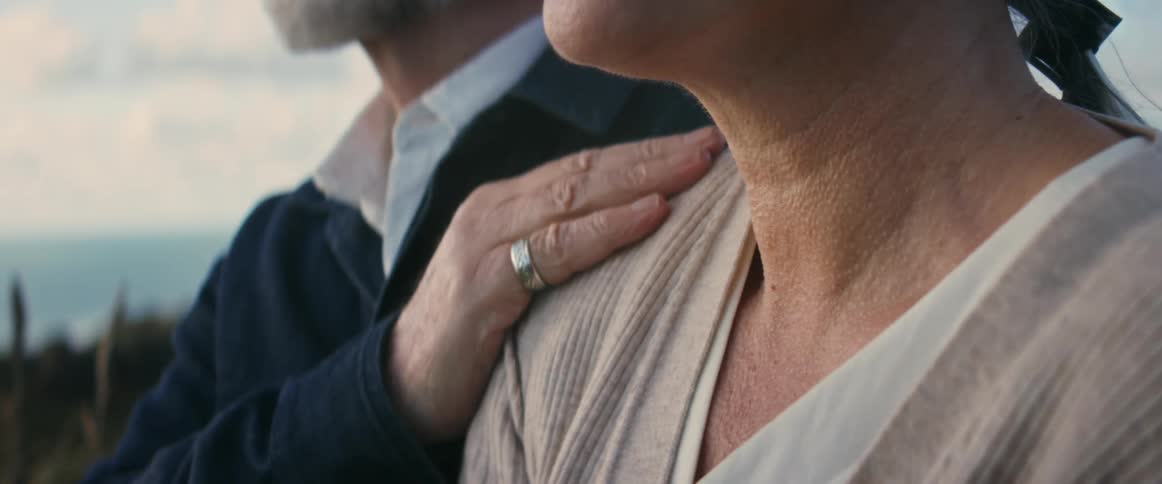 An elderly man stands next to a woman and gently rubs her shoulder