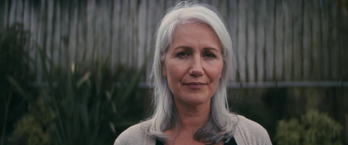 An elderly woman with grey hair staring at the camera intuitively in front of a fence with plants in front of it