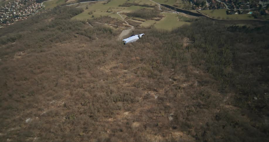 A person wearing a wingsuit jumps off a cliff and flies over trees as they open a parachute and float towards the valley community below