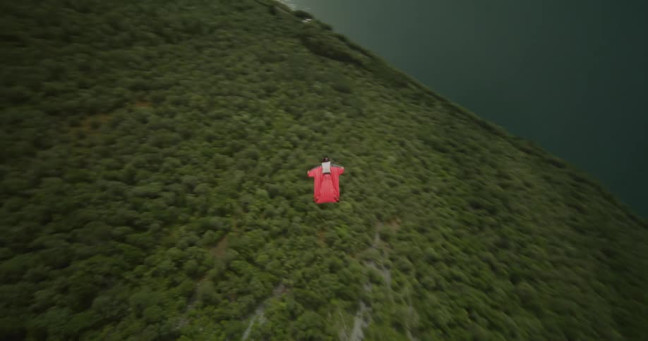 A person wearing a wingsuit is floating along the side of a mountain and flips as they fly toward the river valley