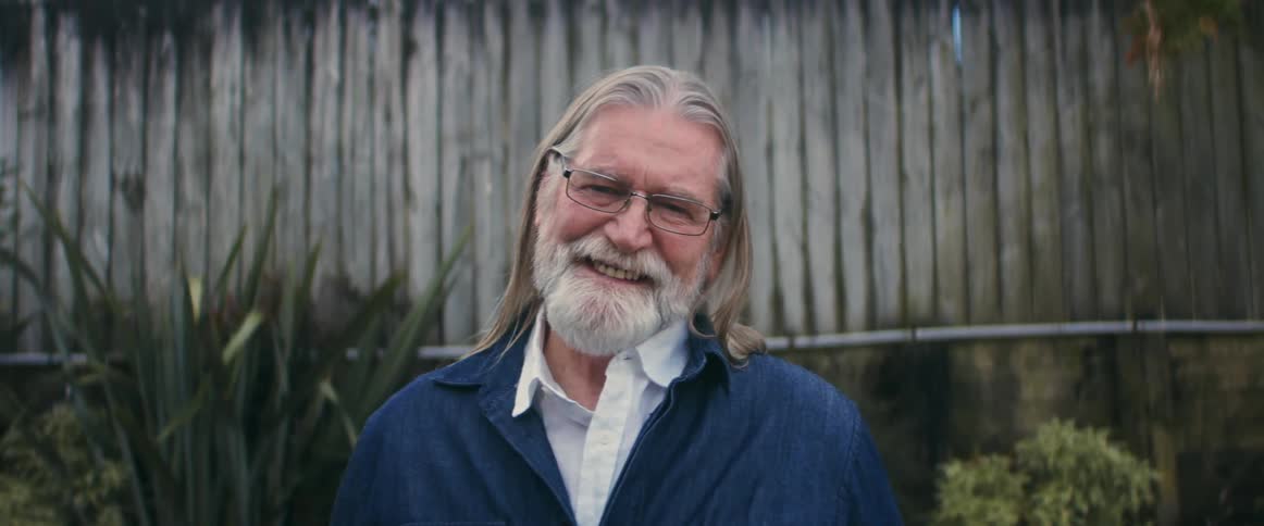 An older man with grey hair and a beard stands laughing and smiling in front of a fence