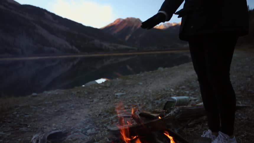 A woman stands warming her hands over a fire near a mountain lake