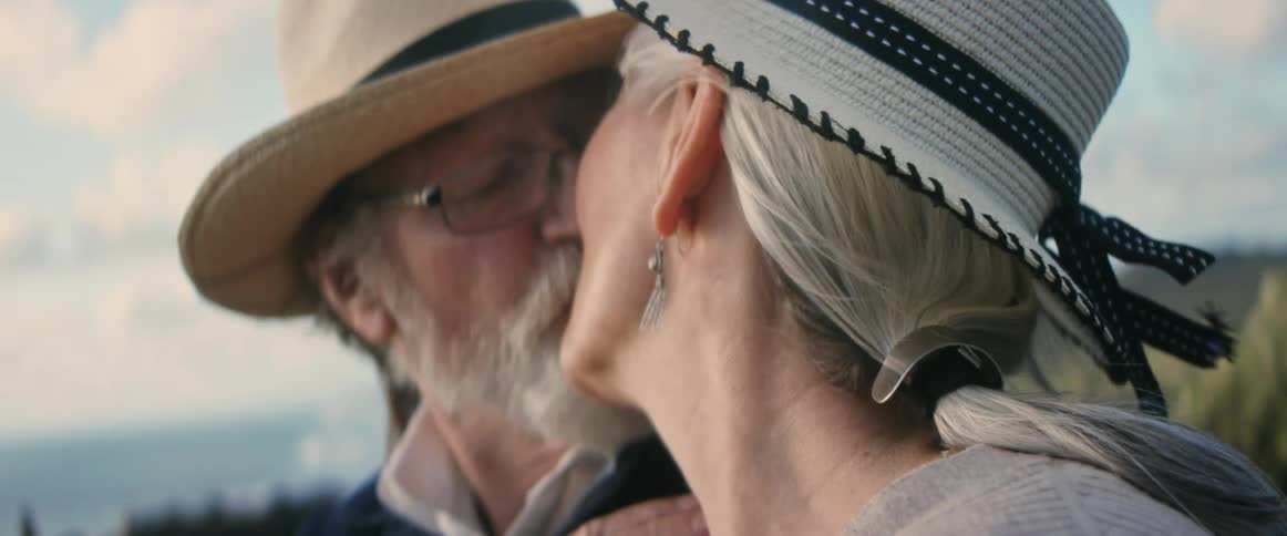 A man and a woman wearing hats are relaxing outside and they look at each other and kiss