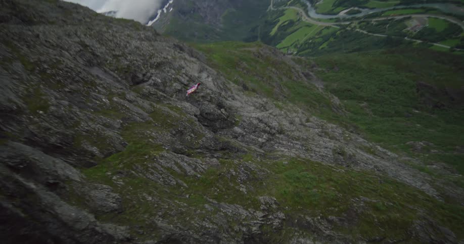 Person wearing winged suit glides near snow dotted mountain towering over valley