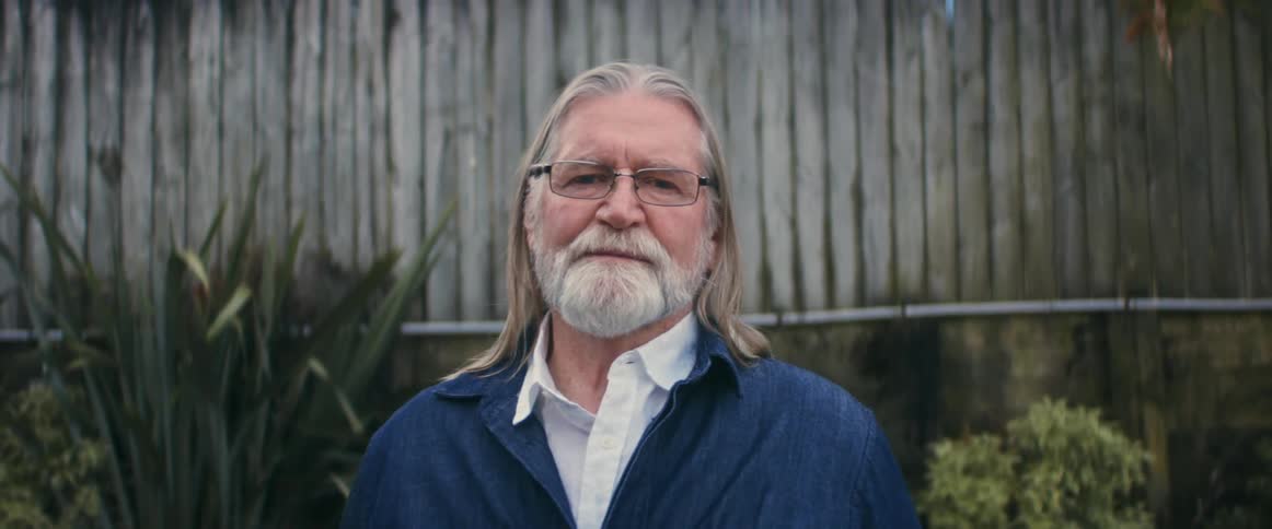 An elderly man is standing in a garden by a fence