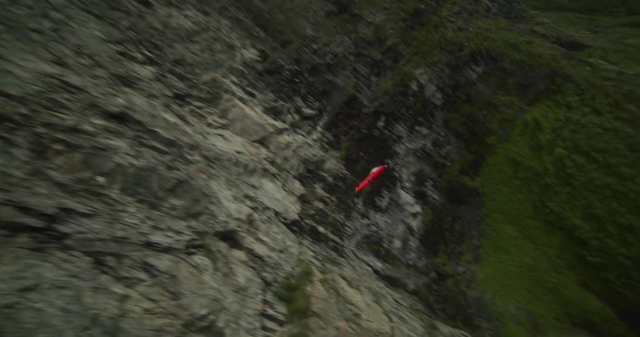 A person wearing a wingsuit jumps off a cliff and tumbles in the air and flips as they float along the side of the cliff and over the valley below