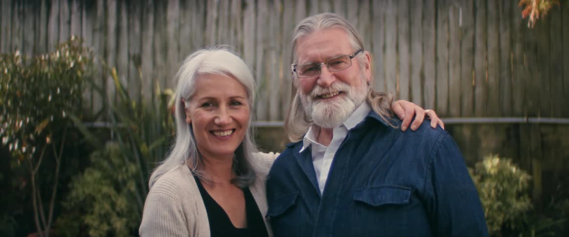 Elderly man with long gray hair glasses and beard and woman with gray hair and brown jacket kiss each other and smiles at camera in the yard