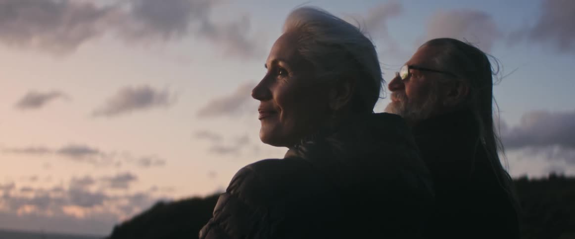 A man and a woman are relaxing along the shore and looking around as the wind blows their hair