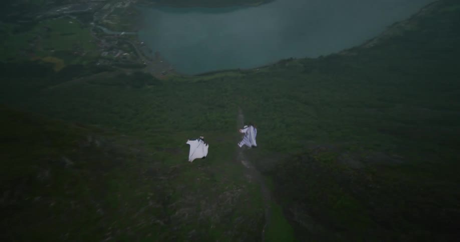 Two people wearing wingsuits jump off a cliff near a building and flip as they fly down towards the lake in the valley below