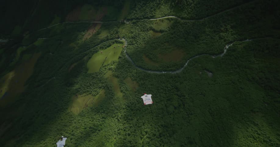 Two people wearing wingsuits are floating over a mountain valley