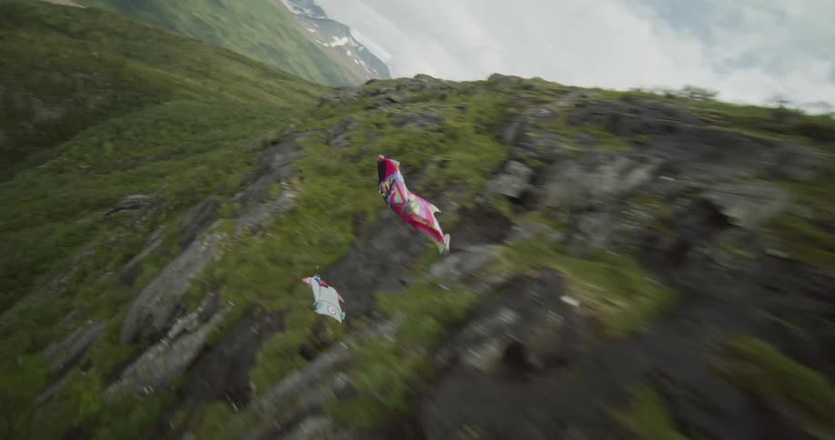 Two people wearing wingsuits are floating along the side of a mountain and flying toward a river valley
